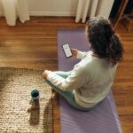 Woman meditating on yoga mat with phone and drink.